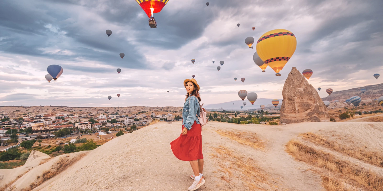 Sunlight aglow on her dress, happy young woman stands in the countryside of Cappadocia, the silhouettes of hot air balloons dancing in the sky above