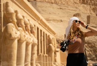 Traveler standing in front of the Mortuary Temple of Hatshepsut in Egypt, wearing sunglasses and a white head covering, holding a camera with colorful strap against a backdrop of sandstone statues and desert cliffs.