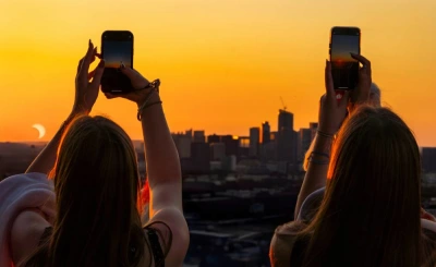 Two travellers photographing the partial phase of a solar eclipse above a city skyline at sunset