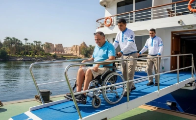   A man in a wheelchair is assisted by two uniformed staff members as he descends a blue ramp from a boat, with palm trees and ancient ruins in the background—showcasing accessible travel in a scenic Egyptian setting.
