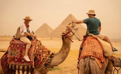 Two tourists riding camels near the Pyramids of Giza, sharing a friendly gesture in the desert haze.