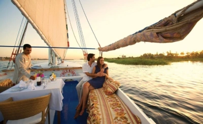 Couple relaxing on a sailboat at sunset, with a server setting a romantic table on calm waters.