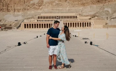 Two visitors stand on a stone path before the Mortuary Temple of Hatshepsut, framed by desert cliffs and colonnaded terraces.