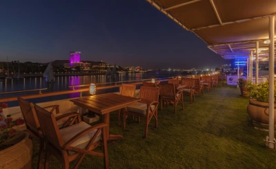 Outdoor terrace dining area at night with lantern-lit tables, river views, and a cityscape illuminated across the water.