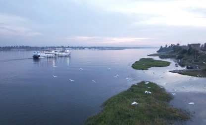 A serene view of a Nile River cruise ship sailing at sunset, with soft pastel skies, calm water, lush green riverbanks, and white birds flying low over the surface.