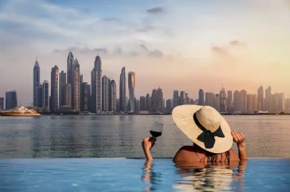 donna con cappello in piedi sul bordo di una piscina e si gode la vista dello skyline della Dubai Marina, Temperature a Dubai donna con cappello in piedi sul bordo di una piscina e si gode la vista dello skyline della Dubai Marina, Temperature a Dubai