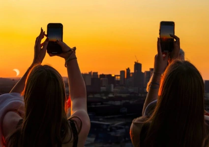 Two travellers photographing the partial phase of a solar eclipse above a city skyline at sunset