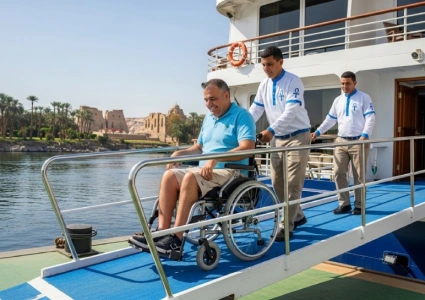   A man in a wheelchair is assisted by two uniformed staff members as he descends a blue ramp from a boat, with palm trees and ancient ruins in the background—showcasing accessible travel in a scenic Egyptian setting.