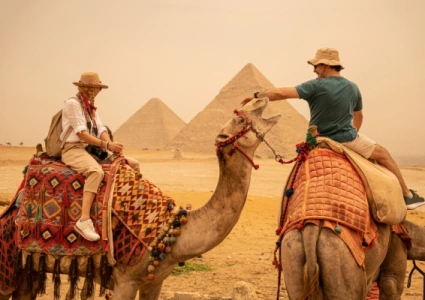 Two tourists riding camels near the Pyramids of Giza, sharing a friendly gesture in the desert haze.