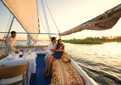 Couple relaxing on a sailboat at sunset, with a server setting a romantic table on calm waters.