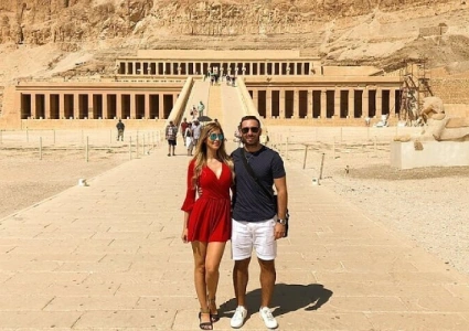 Two tourists standing on the stone pathway leading to Hatshepsut’s Temple, framed by desert cliffs and colonnaded terraces.