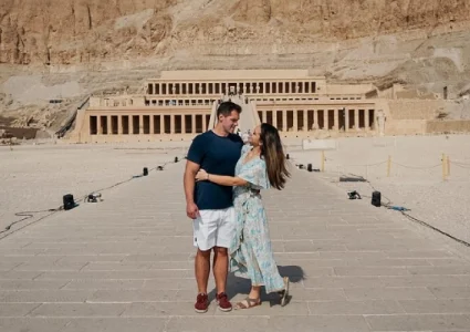 Two visitors stand on a stone path before the Mortuary Temple of Hatshepsut, framed by desert cliffs and colonnaded terraces.