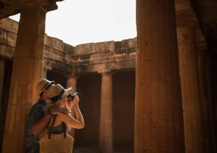 Two tourists exploring an ancient Egyptian temple, one taking a photo amid sunlit stone columns and weathered walls.