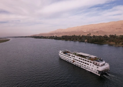 White river cruise ship sailing on a wide river, with lush palm-lined banks and desert hills in the background.