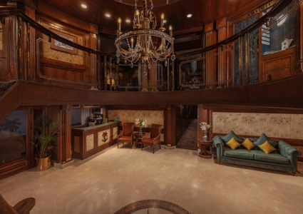 Elegant cruise lobby with a grand chandelier, wood-paneled walls, ornate balcony, and plush seating areas featuring red chairs and a teal tufted sofa.