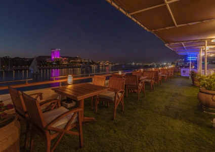Outdoor terrace dining area at night with lantern-lit tables, river views, and a cityscape illuminated across the water.