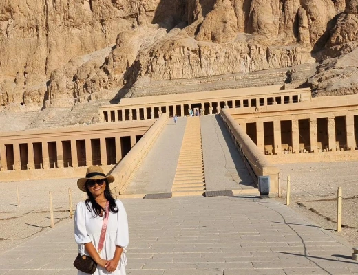 Visitor in white outfit and sunhat standing on pathway leading to the Mortuary Temple of Hatshepsut, built into desert cliffs.