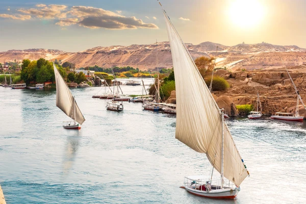 Segelboote (Feluccas) auf dem Nil in Ägypten bei Sonnenuntergang, grünes Ufer links, Wüstenhügel rechts.