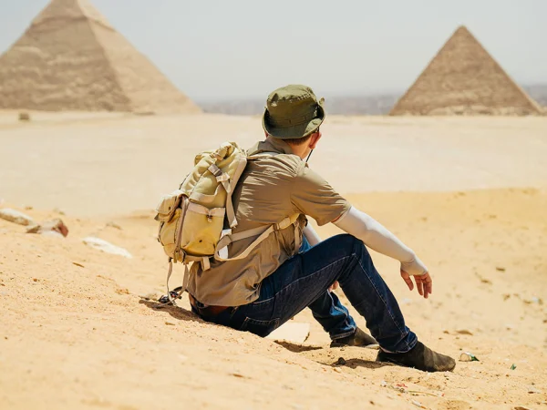 Person sitzt auf einer Sanddüne und blickt auf zwei Pyramiden