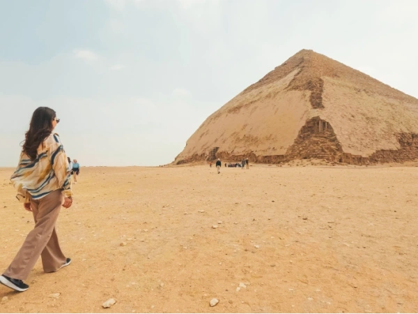Eine Frau geht von hinten gesehen durch eine weite, sandige Wüstenlandschaft auf die Knickpyramide von Dahschur zu, die die rechte Bildseite dominiert.