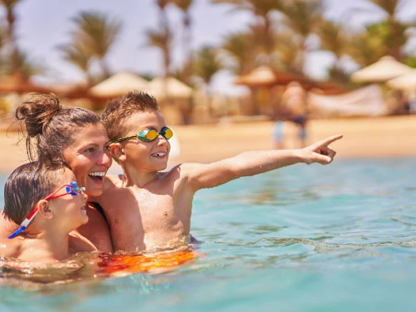 Eine lächelnde Frau und zwei Jungen mit Schwimmbrillen planschen im klaren, türkisfarbenen Wasser eines Strandes; alle blicken nach rechts, wobei der ältere Junge begeistert mit dem Finger in diese Richtung zeigt.