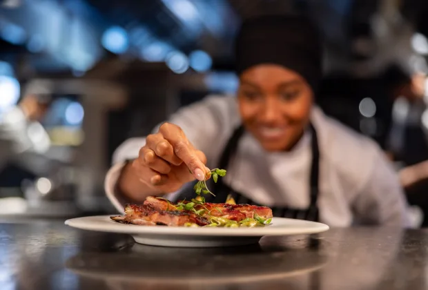 Chef in a professional kitchen carefully placing fresh herbs on a plated dish of cooked meat, with blurred staff and equipment in the background.