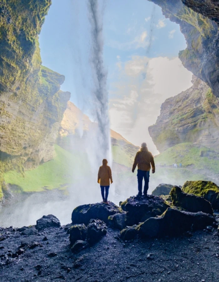Two hikers stand inside a mossy cave, facing a sunlit waterfall cascading from above.