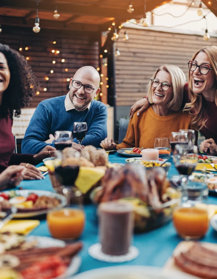 A diverse group of smiling friends and family members enjoy an outdoor dinner party at dusk. They are seated around a long blue table filled with food and drinks, illuminated by warm string lights in the background.