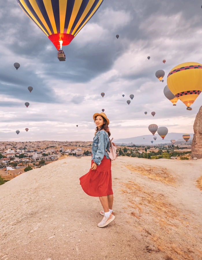 Sunlight aglow on her dress, happy young woman stands in the countryside of Cappadocia, the silhouettes of hot air balloons dancing in the sky above