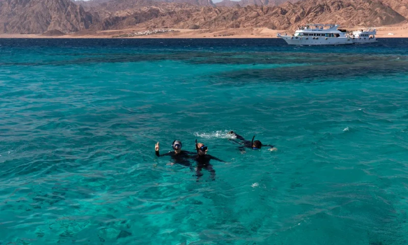 Tres personas haciendo esnórquel en las aguas turquesas del Mar Rojo, con montañas, yates y la ciudad de Dahab al fondo,  Sharm El Sheikh Egipto
