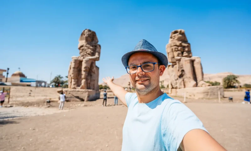Hombre adulto tomándose selfies frente a los Colosos de Memnón en Luxor, Egipto,Colosos de Memnón  .