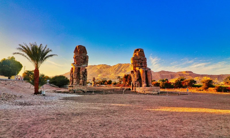 Vista de los Colosos de Memnón en Luxor, Egipto, con las montañas del desierto y un cielo azul despejado al fondo.
