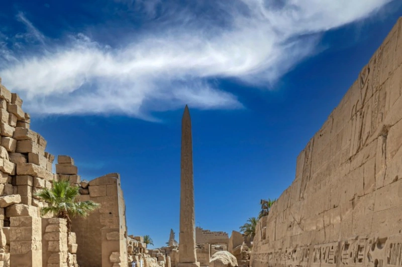 Obeliscos de Egipto en el Templo de Karnak, Luxor, con jeroglíficos tallados en piedra y antiguas estructuras faraónicas bajo un cielo azul.