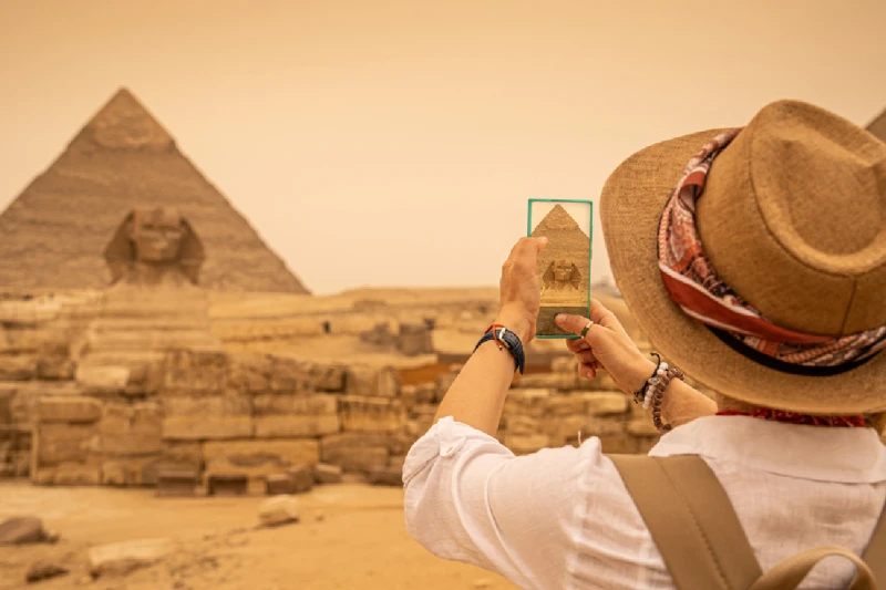 Mujer de pie frente a las pirámides y la Esfinge de Giza, tomando fotos con su smartphone. Giza, El Cairo, Egipto.