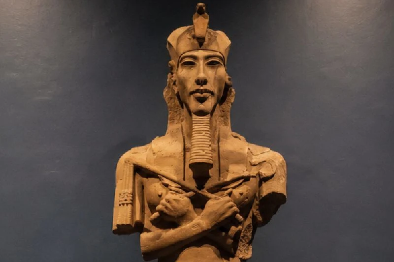 Estatua completa del faraón Ajenatón expuesta en un museo, iluminada sobre un fondo oscuro y centrada en la imagen.