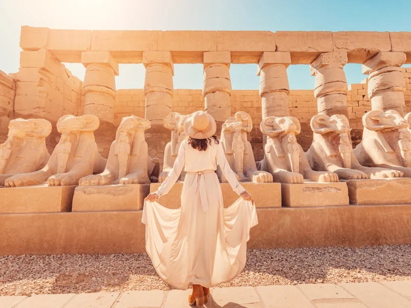 Voyageuse en robe blanche devant l'allée des sphinx à tête de bélier du temple de Karnak à Louxor sous le soleil de décembre