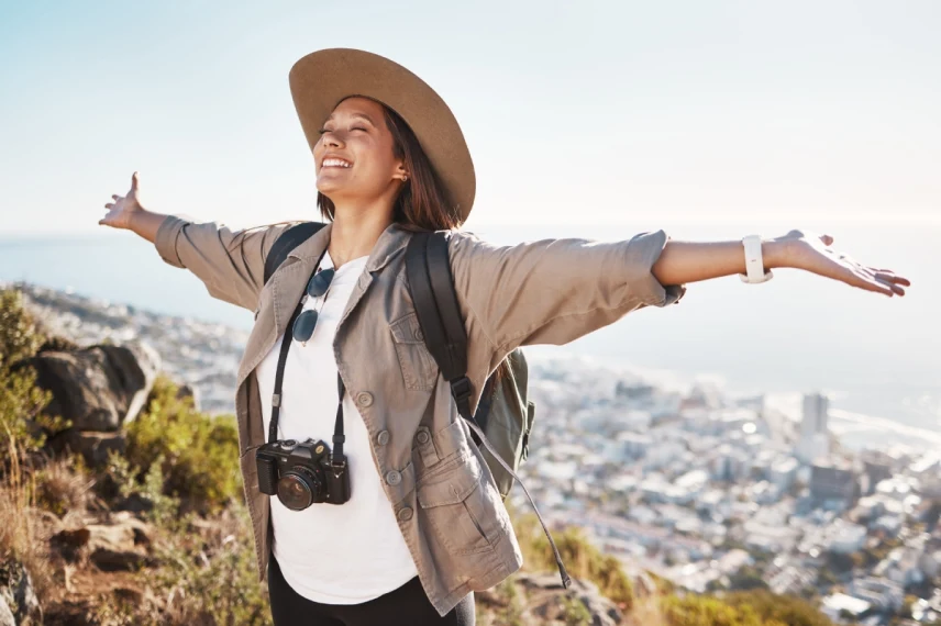 Traveler standing on hillside, arms outstretched, overlooking coastal city and ocean.