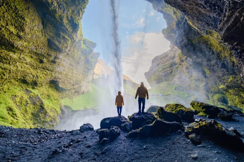 Two hikers stand inside a mossy cave, facing a sunlit waterfall cascading from above.