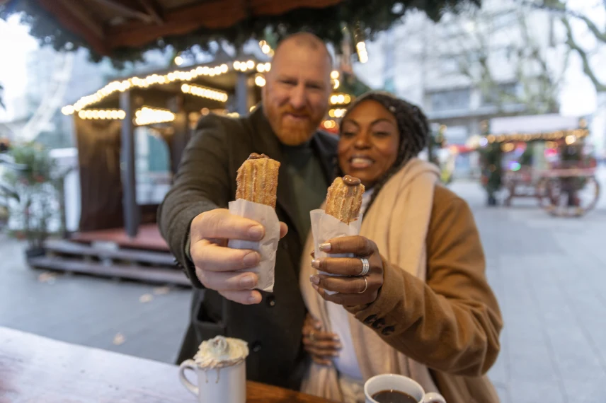 Two people holding churros with chocolate sauce at a festive outdoor market, with mugs of hot drinks on a wooden counter.