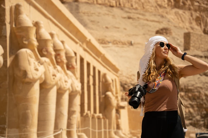 Traveler standing in front of the Mortuary Temple of Hatshepsut in Egypt, wearing sunglasses and a white head covering, holding a camera with colorful strap against a backdrop of sandstone statues and desert cliffs.