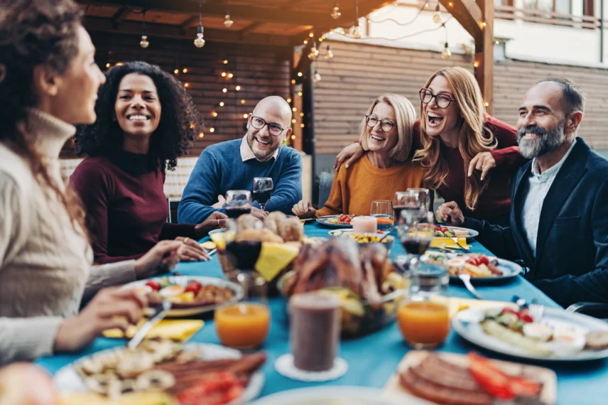 A diverse group of smiling friends and family members enjoy an outdoor dinner party at dusk. They are seated around a long blue table filled with food and drinks, illuminated by warm string lights in the background.