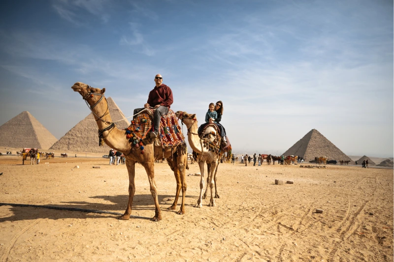 Viajar a Egipto 2025 Dos personas montando camellos en un vasto paisaje desértico bajo un cielo azul claro, con mantas de silla de montar tradicionales y un horizonte lejano.