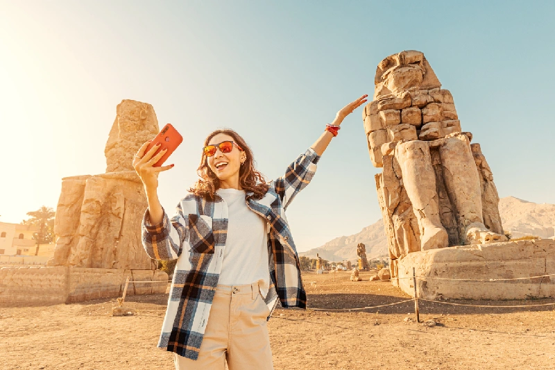los famosos dos Colosos de Memnón Una chica se toma selfies con su teléfono con los famosos dos Colosos de Memnón, enormes estatuas en ruinas del faraón Amenhotep III. Atracciones turísticas mundiales.