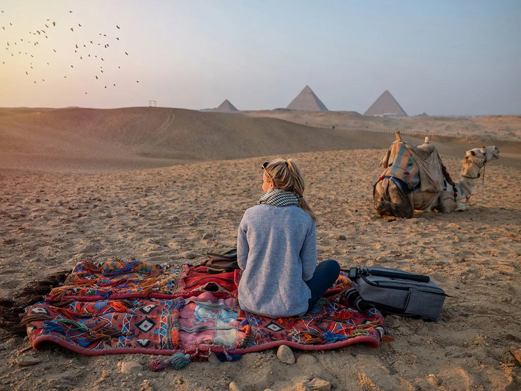 a blond girl sitting infront of pyramids of Giza and a camel beside her