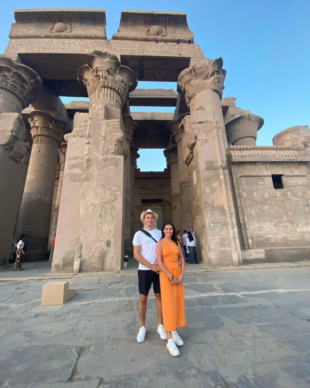 Two people standing before the towering stone columns of the Temple of Kom Ombo, carved with hieroglyphs and reliefs.