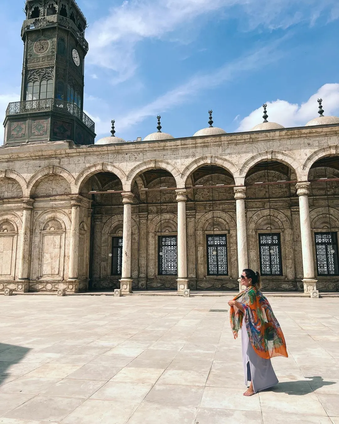 Person in colorful shawl standing in courtyard with ornate arches, domes, and a tall clock tower showing Islamic architectural design.
