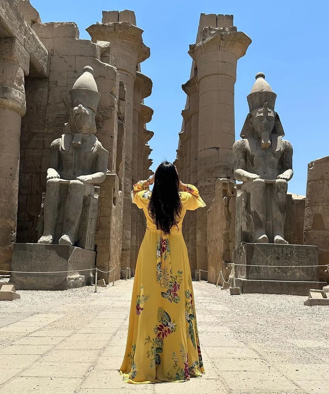 Person in bright yellow floral dress facing massive seated pharaoh statues at temple entrance, with tall stone columns under clear sky.