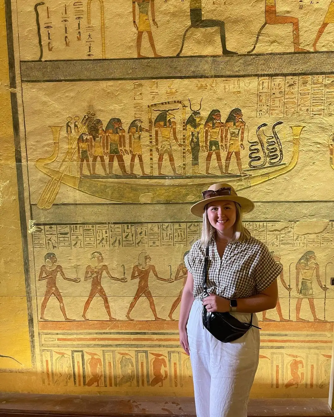 Visitor in hat and checkered shirt smiling before ancient Egyptian wall mural with hieroglyphs and deities, including figures on a boat.