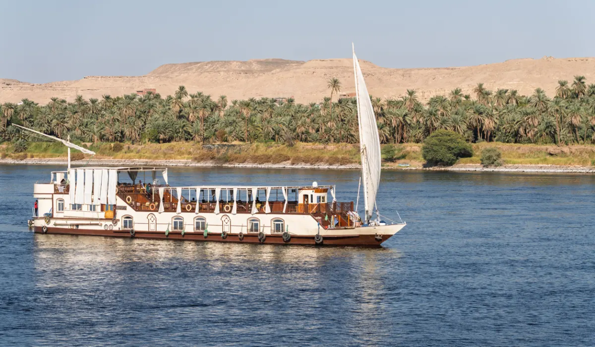 Dahabiya Nile cruise ship sailing alongside a sandy desert with palm trees on the River Nile near Aswan