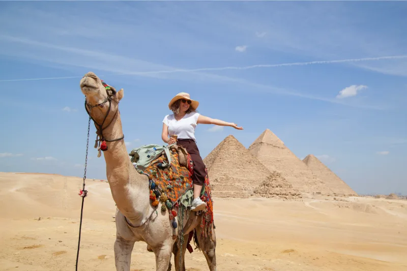 Una turista montando un camello detrás de ella se encuentran las grandes pirámides de Giza